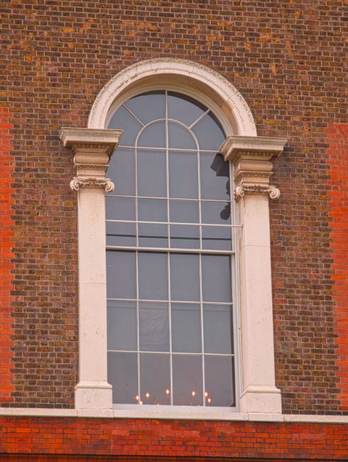 The exterior of a red brick residential building with four white-framed sash windows, each featuring a rectangular windowpane arrangement. The two upper windows are adorned with window boxes containin