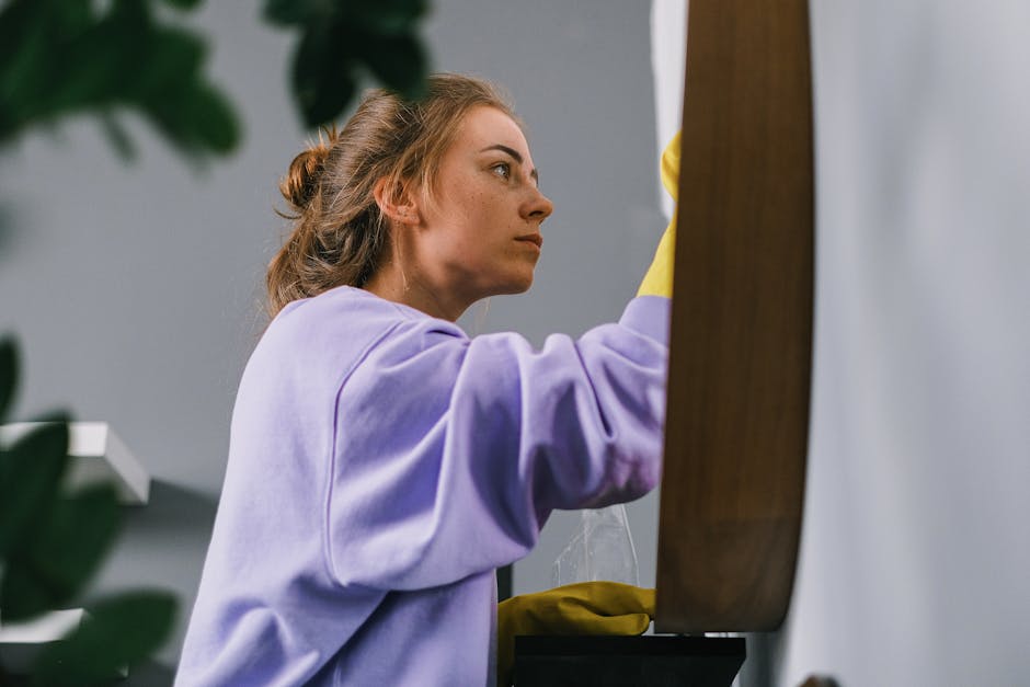 A young woman with light brown hair tied back, wearing a lavender sweatshirt and yellow cleaning gloves, focused on cleaning a wooden surface with a cloth. The background features a neutral-colored wa